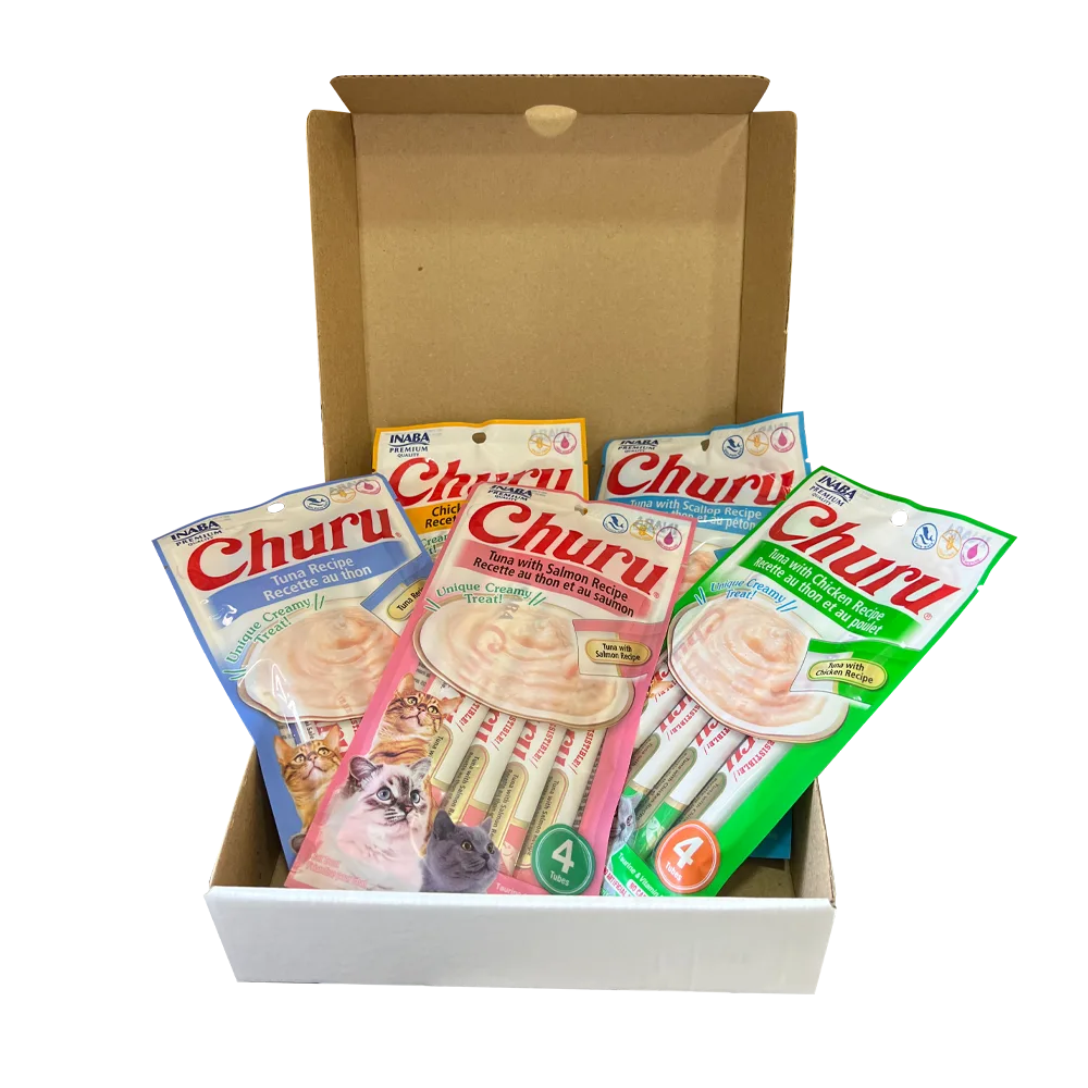 Caja navideña de cartón colo blanco con un gato gris en la cara frontal, a su lado hay 4 empaques de los Snacks Churu en sus distintos sabores y un plato de goma "Lick Mat" azul