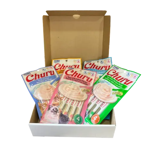 Caja navideña de cartón colo blanco con un gato gris en la cara frontal, a su lado hay 4 empaques de los Snacks Churu en sus distintos sabores y un plato de goma "Lick Mat" azul